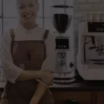 Female barista standing infront of a cafe bench top, where a Brasilia commercial coffee machine and grinder sit